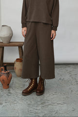 A woman stands in a studio wearing a matching brown two-piece outfit consisting of a long-sleeve collared top and wide-leg cropped pants, paired with brown lace-up boots. The background features a light gray floor, a white backdrop, and a small wooden stool holding a ceramic vase.