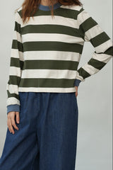 A person wearing a long-sleeve striped knit top with horizontal olive green and off-white stripes, paired with loose blue denim pants, against a plain light gray studio background.