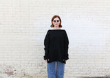 A woman with short auburn hair and sunglasses wearing a loose black long-sleeve top and blue jeans, standing against a white brick wall.