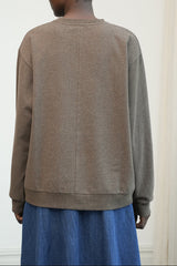 A person shown from the back wearing a taupe-brown crewneck sweatshirt and a blue skirt, standing against a light off-white wall.
