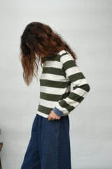 A woman with long brown hair is shown from the side, wearing a green and off-white horizontally striped long-sleeve top with a blue collar and dark blue wide-leg pants, set against a plain light gray background.
