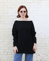 A woman with red hair and sunglasses wearing a black off-the-shoulder long-sleeve top and blue jeans, standing against a white brick wall.