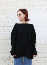 A woman with short auburn hair and glasses wearing a loose black off-the-shoulder long-sleeve top and blue jeans, standing against a white painted brick wall.